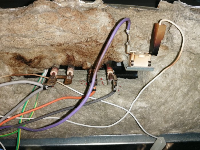 Gamla elektriska komponenter och kablar mot isoleringsmaterial i en dammig, oordnad installation. Säkerhetsrisk synlig.