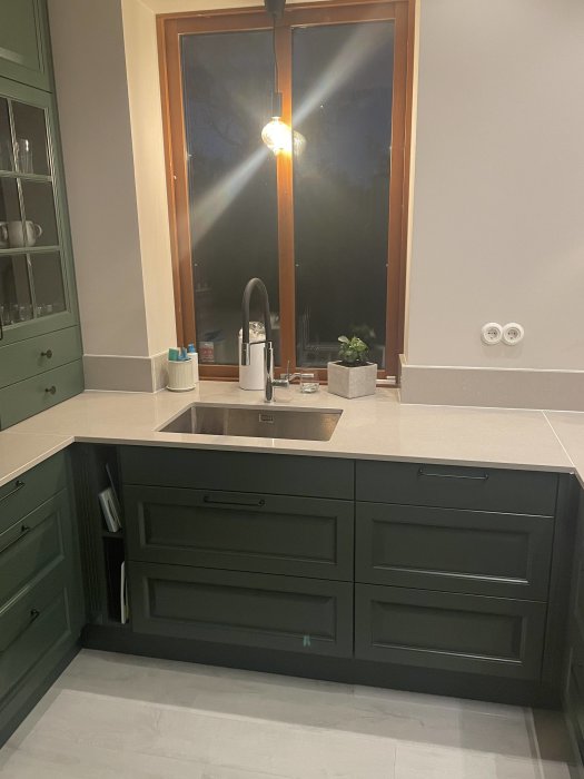 Köksinteriör med mörkgröna skåp, diskbänk, spegel och fönster vid skymningen. Modern och stilren design.