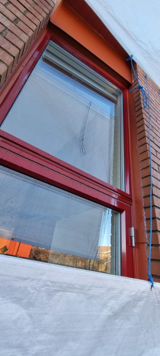 Ett fönster med röda kanter, skyddat av presenning, blått rep och tegelvägg ovanför.