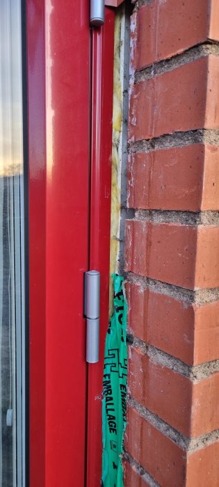 Röd dörrkant, isoleringsskum, tegelvägg, grön plastduk som tätning, solljus.