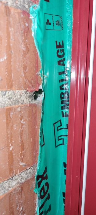 Grön byggfolie nära rött fönster och tegelvägg, skadad isolering, läckage eller pågående konstruktion.