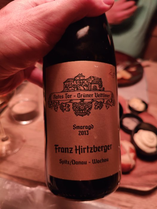 En hand håller en vinflaska med etikett "Rotes Tor - Grüner Veltliner, 2013, Franz Hirtzberger, Wachau". Suddig bakgrund, maträtter.