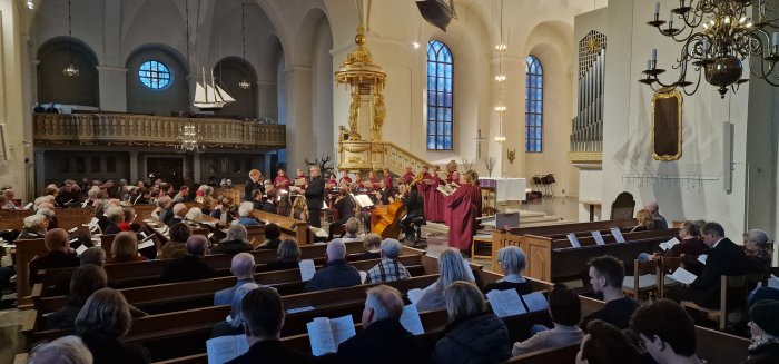 Kör framför publik i kyrka, musiker, dirigent, stämningsfull belysning, konserthändelse, arkitektoniska detaljer, avslappnad atmosfär.