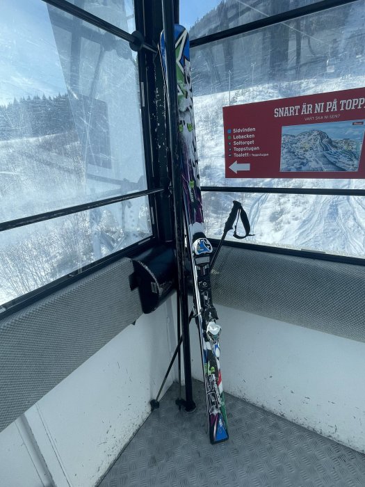 Skidor och stavar stående inne i en skidliftgondol, snöigt bergslandskap synligt utanför, klar himmel.