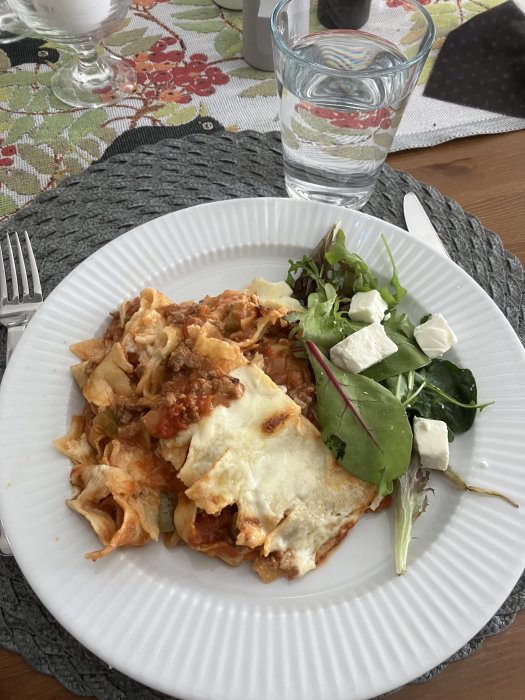 Lasagne på tallrik med sallad, fetaost, vattenglas och bestick på matbord.