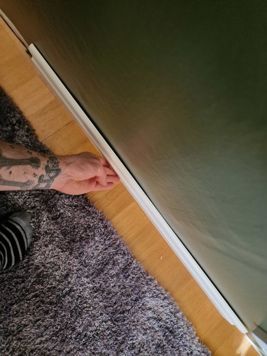 En person som ligger på golv tar i en dörrkant, tatuering synlig, randig socka, lila matta.