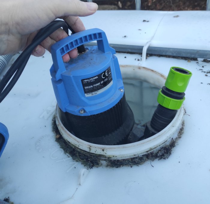 Hand lyfter blå dräneringspump ur vattentank med grön slangkoppling bredvid. Smutsiga detaljer, utomhus.