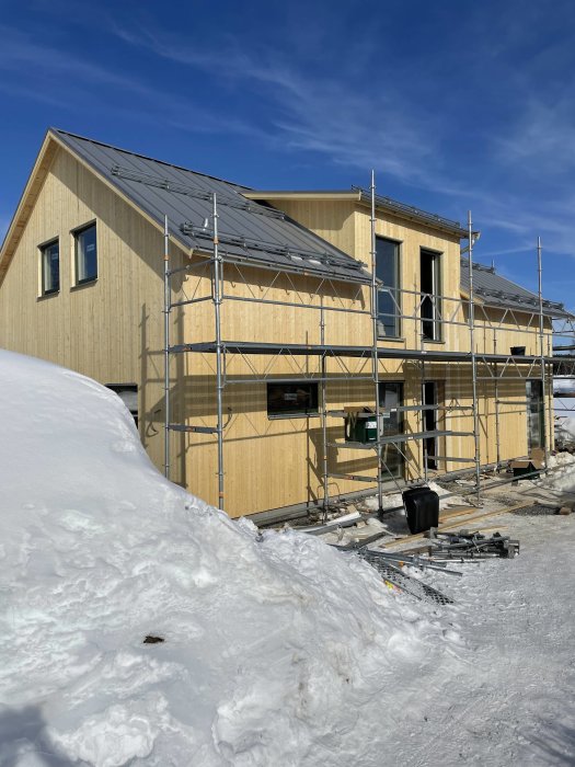 Nybyggd trähus under konstruktion med ställningar, omgiven av snö, klart väder.