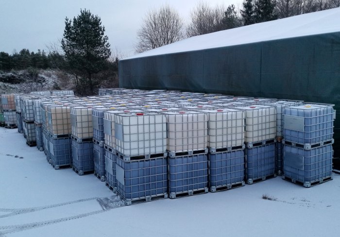 Staplar av IBC-containrar utomhus i snö, lagring, industri, träd, grå himmel, vintertid.