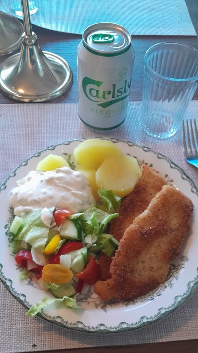 Ett måltidsarrangemang: stekt fisk, potatisar, sallad, sås, ölburk och vattenglas på dukat bord.