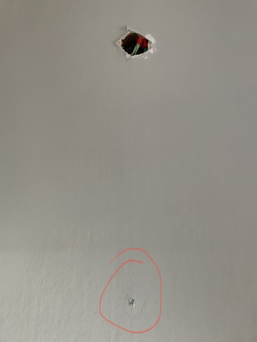 Gråmålad vägg med hål och kablar synliga, röd cirkel markerar mindre skada.