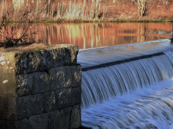 Lugnt vattenfall, stenvägg, återspegling av träd, kvällsljus, orörd natur.