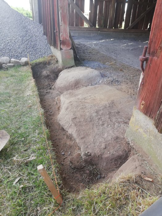 Utgrävning nära träbyggnad, stora stenar, grus, snöre spänner, trästolpe, gräs, grävverktyg syns ej. Konstruktion eller anläggningsarbete antyds.