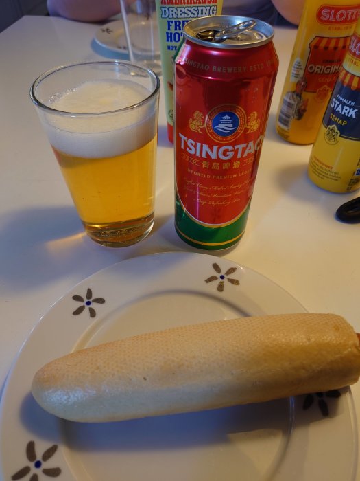 Ett glas öl, en Tsingtao-ölburk och en korv på en tallrik.