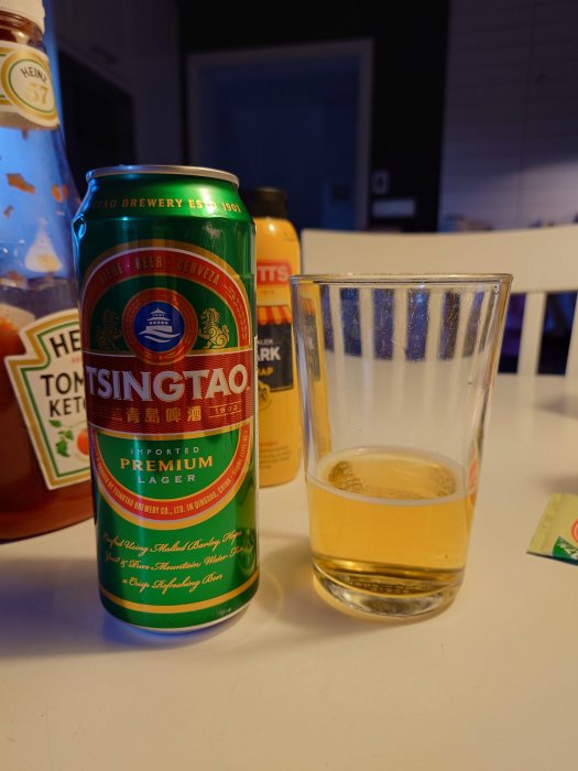 Tsingtao-ölburk bredvid öppet glas av öl, ketchup och sås på bord i bakgrunden.