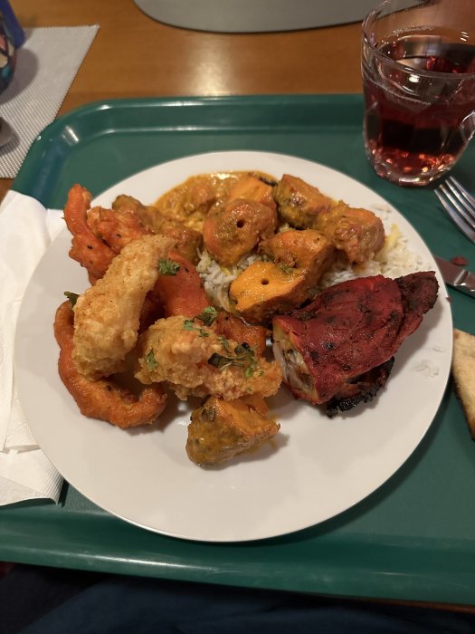 Tallrik med blandade maträtter inklusive friterade räkor, grillat kött och curry, serverad med ris och dryck.
