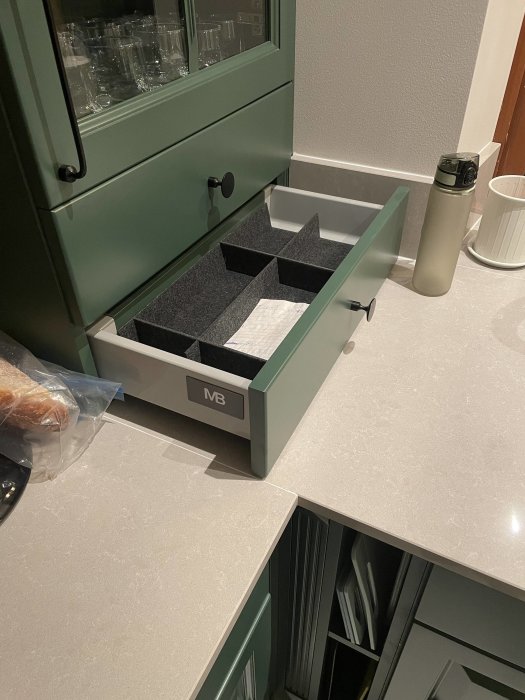 Ett grönt köksskåp är öppet, visar en välorganiserad låda med fack, nära en stengrå bänkskiva.