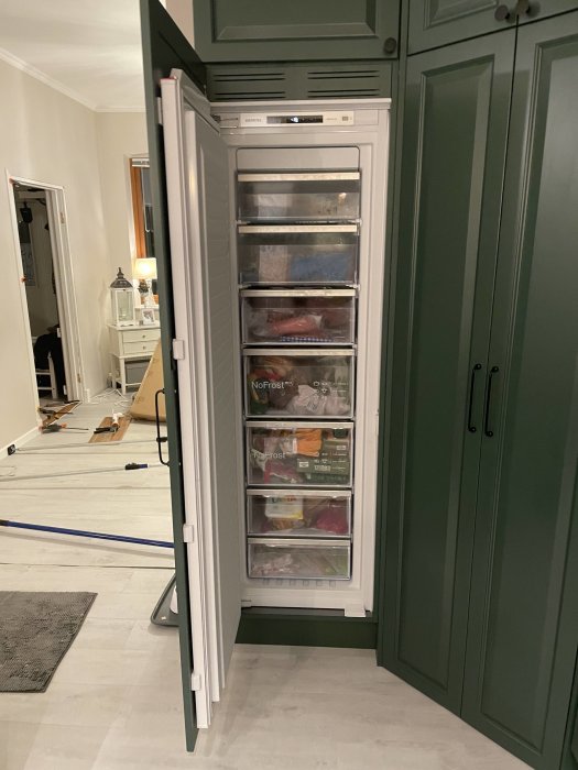 Ett öppet frysskåp med lådor, i ett kök under renovering, verktyg synliga på golvet.