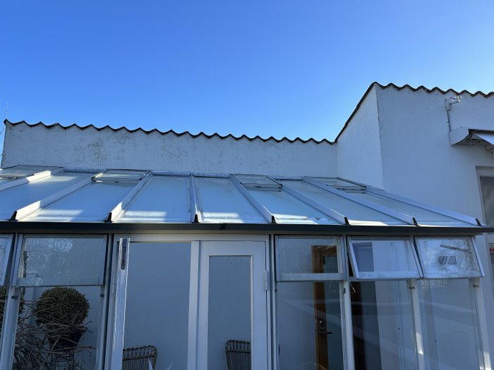 Glasveranda under klarblå himmel, vit vägg med mönster, tegel och antenner syns också.