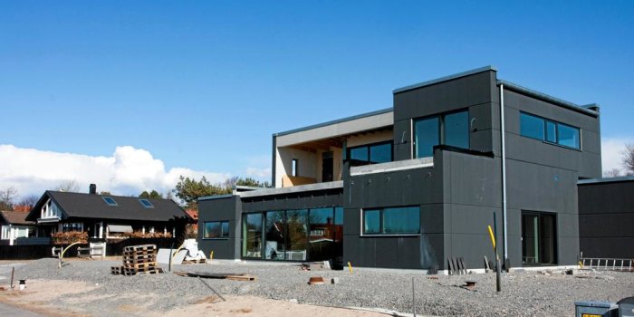 Modernt hus under konstruktion med byggmaterial framför, traditionella hus i bakgrunden och klarblå himmel.