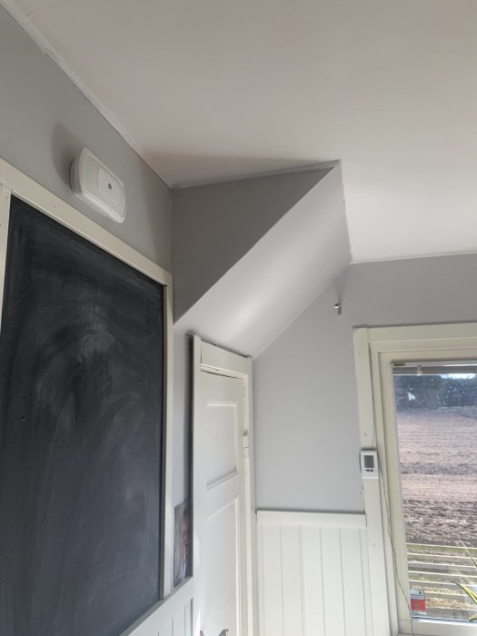 Ett rumshörn med brandvarnare, svarta tavlan, dörr och fönster som visar utsikt av fält.