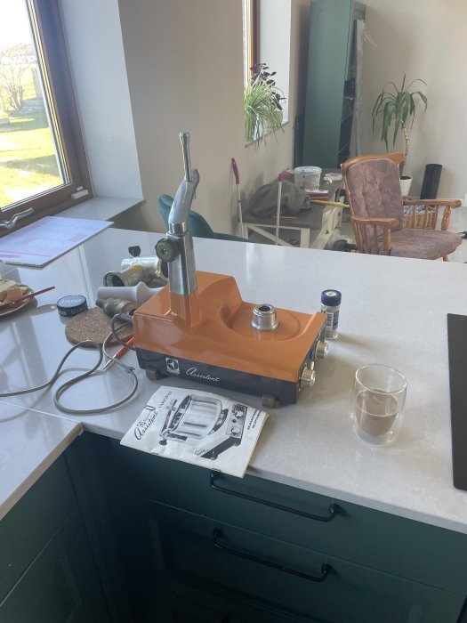 Espressomaskin på köksbänk, glas med dryck, manual, inomhusmiljö med fönster och möbler i bakgrunden.