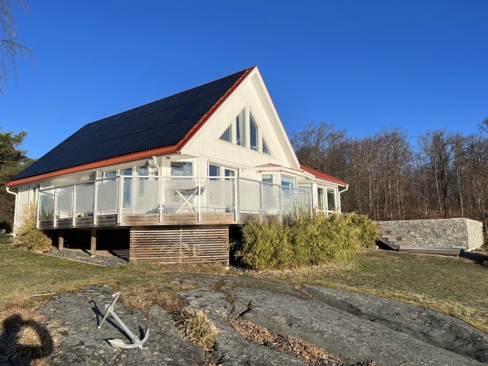 Vitt hus med glasveranda, solpaneler på taket, ankardekoration, stenmur och trädäck på klippa, klart väder.