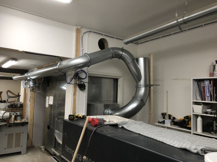 Verkstadsinteriör med ventilationssystem, maskiner, verktyg och arbetsbänk. Industriell atmosfär, pågående installation eller underhåll.