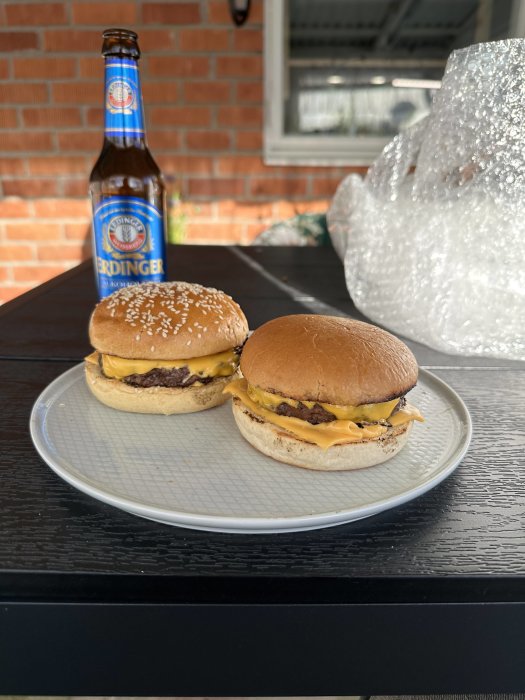 Två ostburgare på tallrik, en ölflaska, utomhus på ett bord, bubbelplast i bakgrunden.