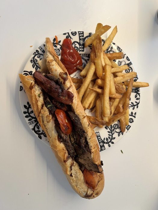 En baguette med kött och tomat, pommes frites och ketchup på ett mönstrat fat.