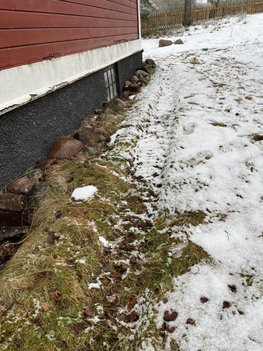 Smältande snö vid husvägg, spår i gräs, stenrader, brun-röd byggnad, gråmulet väder, vinter till vår övergång.