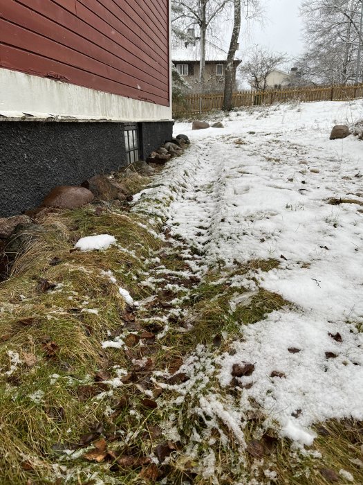 Snötäckt gräsmatta bredvid en röd trähusvägg, stenar, smältande snö, nakna träd, gråmulen himmel.