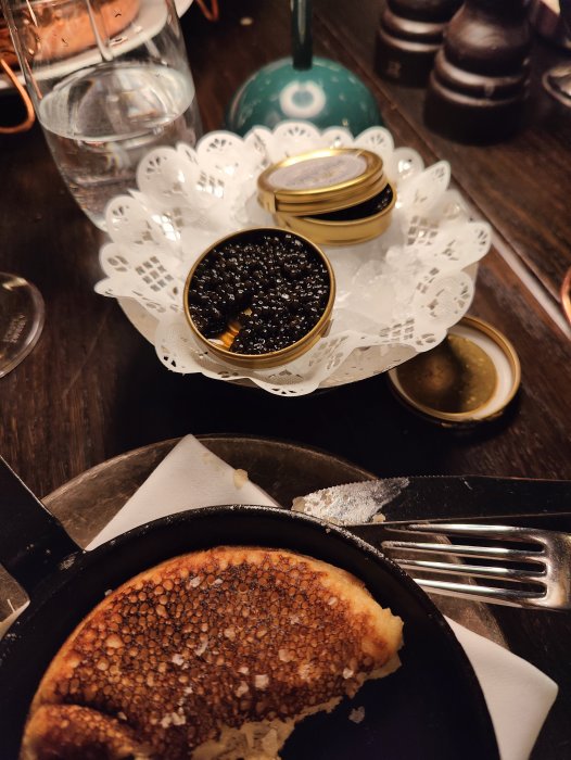 Lyxig måltid med kaviar i burk, blini, bestick och glas vatten på träbord.