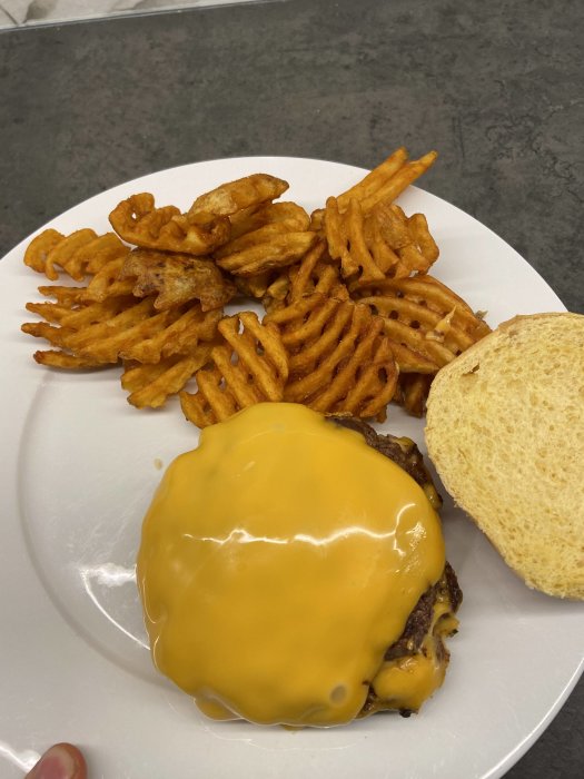 Cheeseburger med smält ost, waffle fries, en bit bröd, på en vit tallrik.