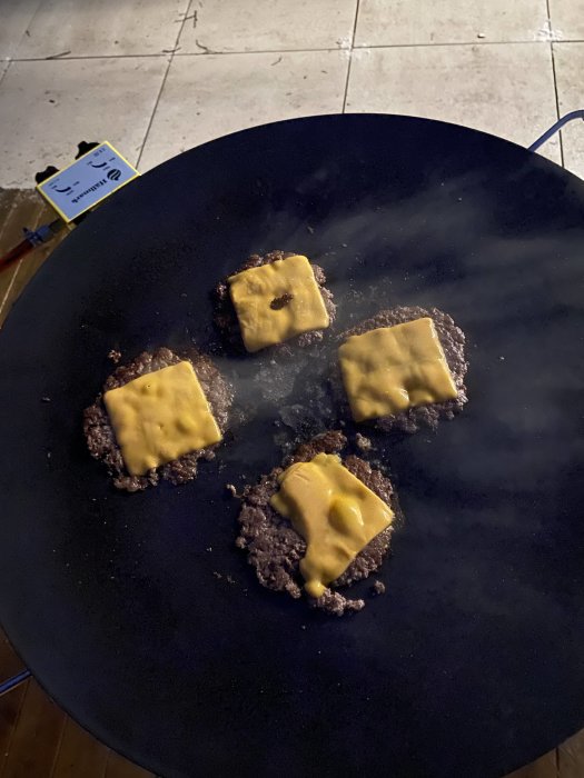 Fyra hamburgare med smält ost steks på stor svart stekyta utomhus i kvällsljus.