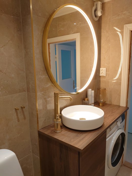 Modernt badrum, trätema, rund spegel, guld detaljer, vita väggar, tvättmaskin, integrerad handfat på arbetsbänk.