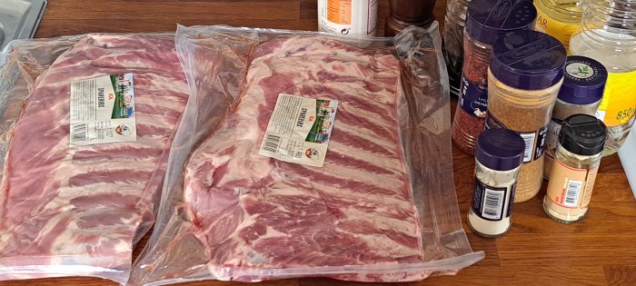 Två förpackningar råa spareribs och olika kryddor på ett träbord, förmodligen förberedelse för matlagning.