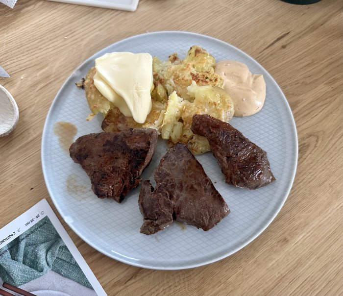 Middagstallrik med stekt kött, potatisklyftor, smörklick och sås. Enkel hemlagad måltid, träbord i bakgrunden.