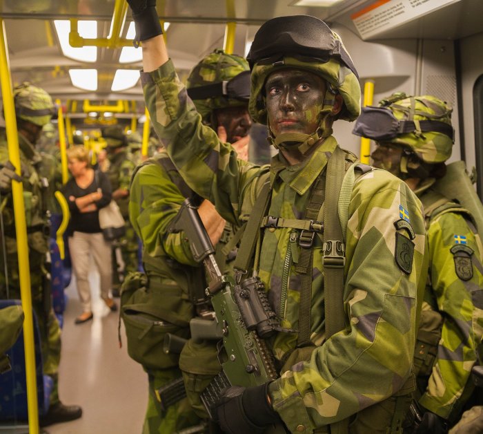 Soldater i uniform med kamouflagefärg i ansiktet, vapen, på ett tåg, civilpersoner i bakgrunden.