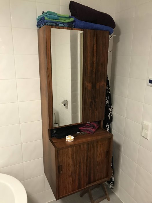 Ett badrumsskåp med spegel, handdukar uppepå och föremål inne i skåpet och på hyllan.