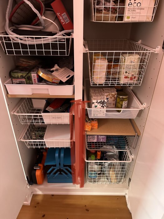 Ett öppet förvaringsskåp fullt av hyllor och korgar med diverse föremål och hushållsprodukter, organiserat i en korridor.