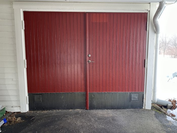 Röda garageportar med svart nederdel på en vit byggnad, snö synlig, fuktig asfalt.
