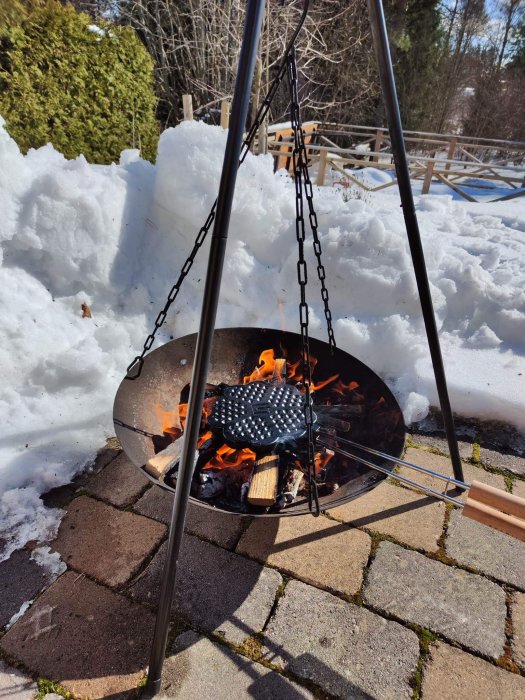 Eldstad utomhus med kokkärl ovanför öppen eld, omgiven av snö, soligt vinterdag.