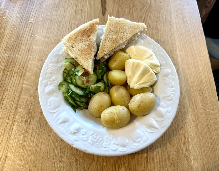 En tallrik med potatis, mackor, gurksallad och klickar av smör på ett träbord.