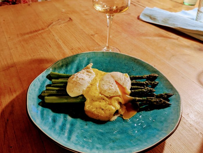 Sparris med pocherade ägg och hollandaisesås på blå tallrik, vin i glas, träbord i bakgrunden.