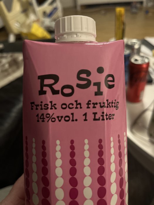 Rosa dryckesförpackning märkt "RoSie", 14% alkoholvolym, 1 liter, punktmönster, otydlig bakgrund.