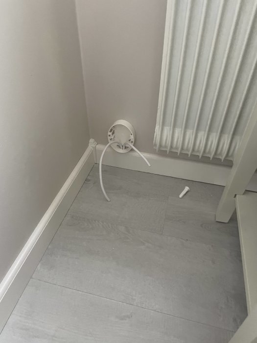 Vit hörna i rum med vägguttag, radiator, grått golv, vit kabel och plastbit.