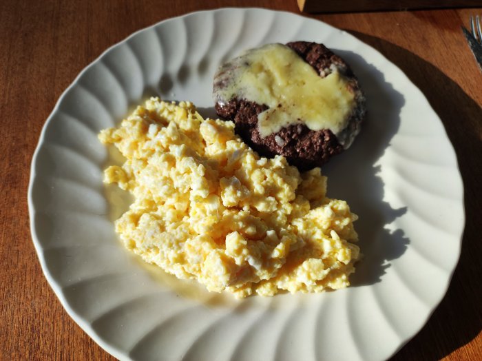 Tallrik med äggröra och köttbulle med ost, träbord, skuggor, solljus, frukost eller lunch.