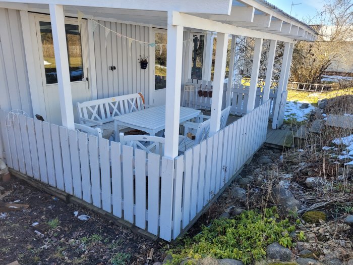 Vit veranda med staket, utemöbler, snörester, och grönska utanför ett hus i solljus.