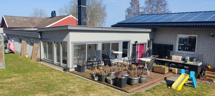 Utomhus, hus med terrass, solpaneler, leksaker, trädgårdsmöbler, planta, barnrutschkana, klart väder.